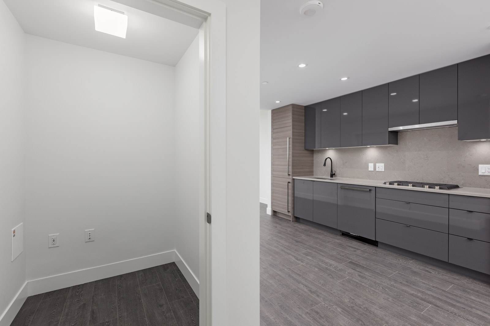 Modern kitchen with gray cabinets and appliances on the right, adjacent to a small empty room on the left. Dark wood flooring throughout.