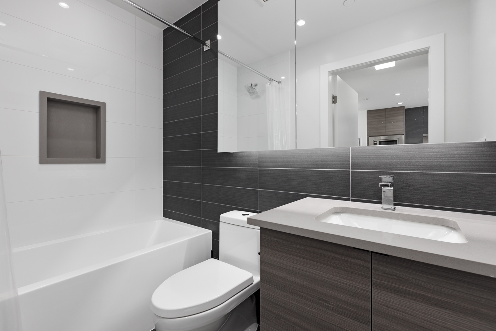A modern bathroom with a white bathtub, toilet, and a sink on a wooden vanity. The walls are tiled in white and dark gray, and a large mirror hangs above the sink.