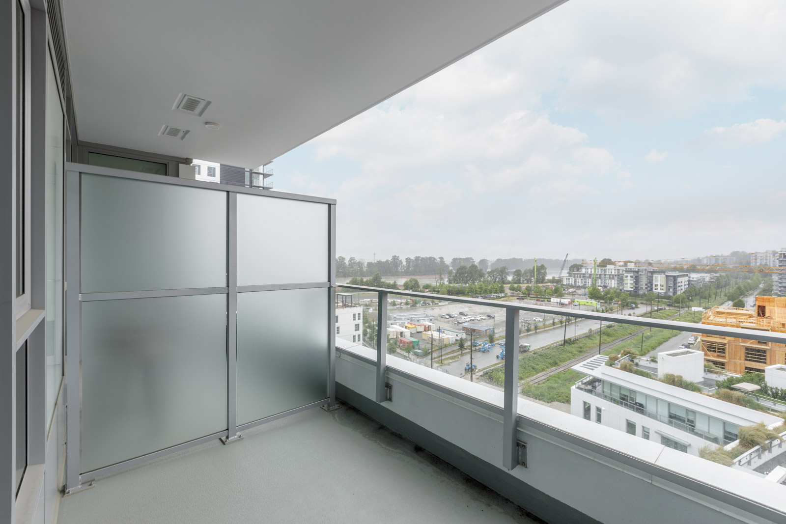 Apartment balcony with frosted glass partition and railing overlooking a residential area and cloudy sky.