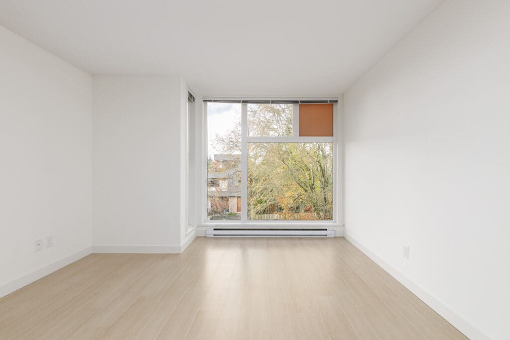 Empty room with light wood flooring, white walls, and a large window showing trees outside.