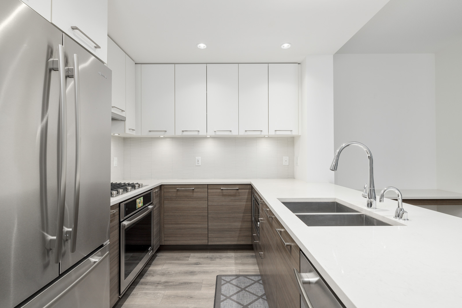 Modern kitchen with stainless steel appliances, white cabinets, and wooden lower cabinets. Double sink and sleek countertops on a light wooden floor.