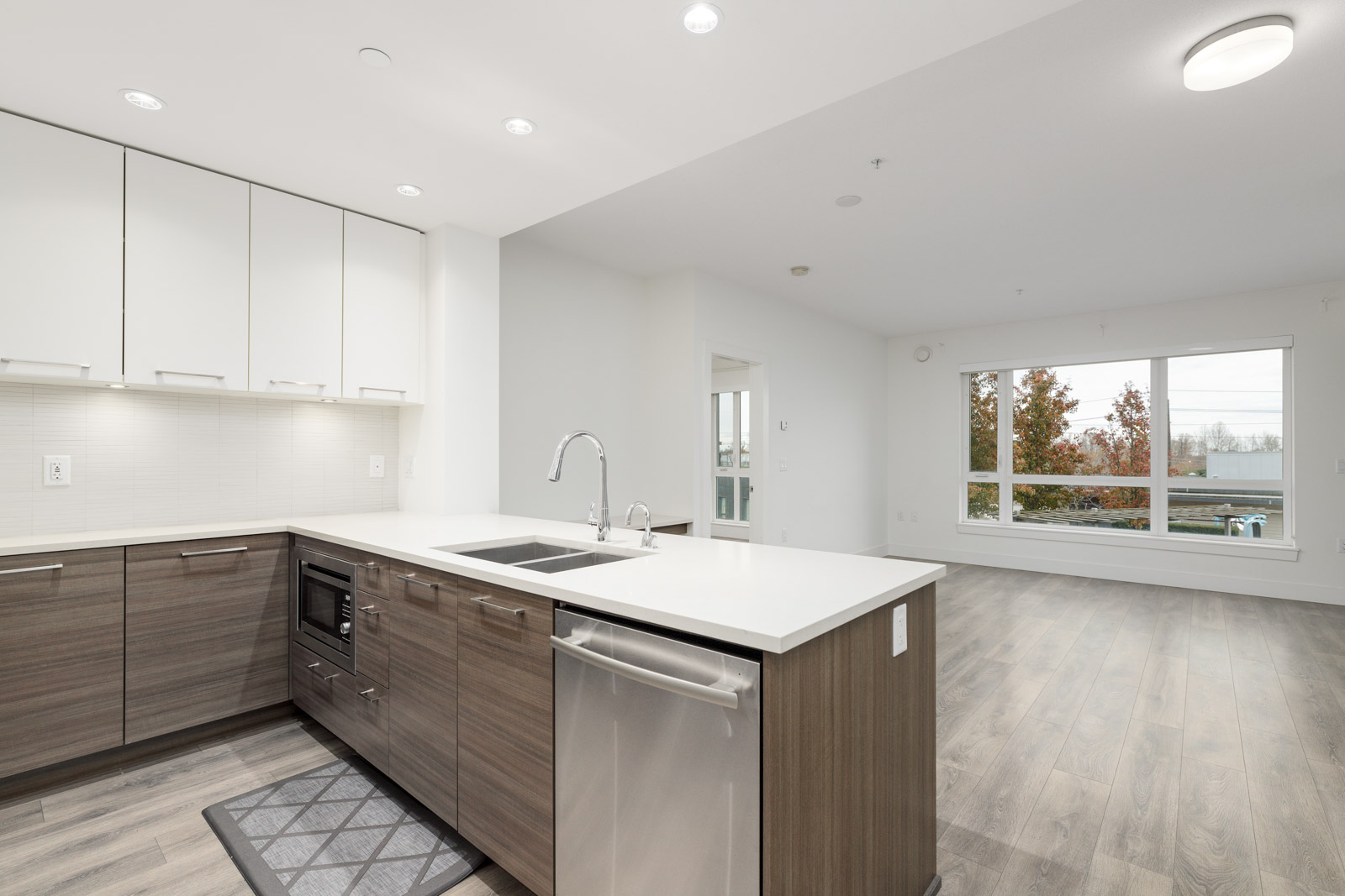 Modern kitchen with white and wood cabinets, an island with a sink, and stainless steel appliances; open layout leads to a living room with large windows and hardwood flooring.