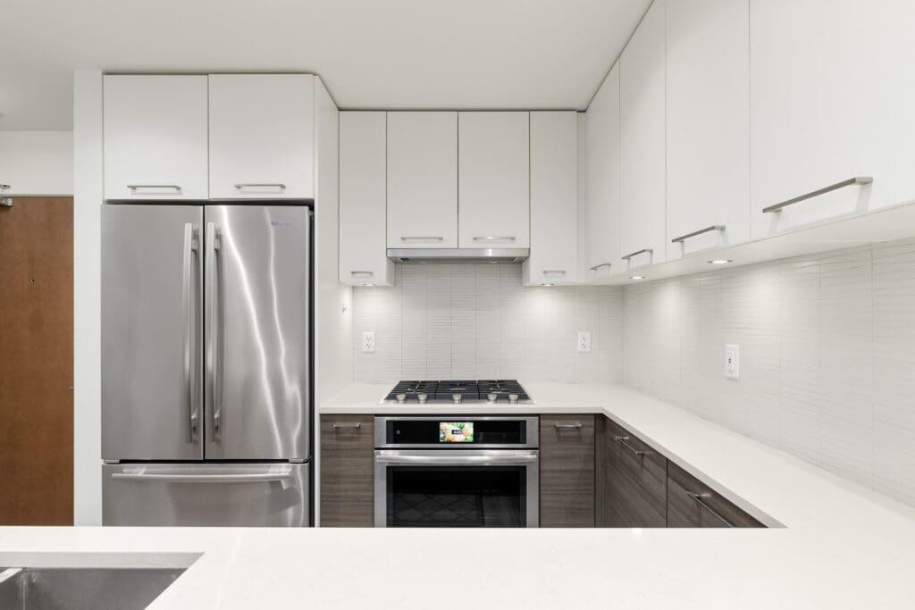 Modern kitchen with white cabinets, stainless steel refrigerator, stove, and oven. The countertops are white, and the backsplash is light grey tile.