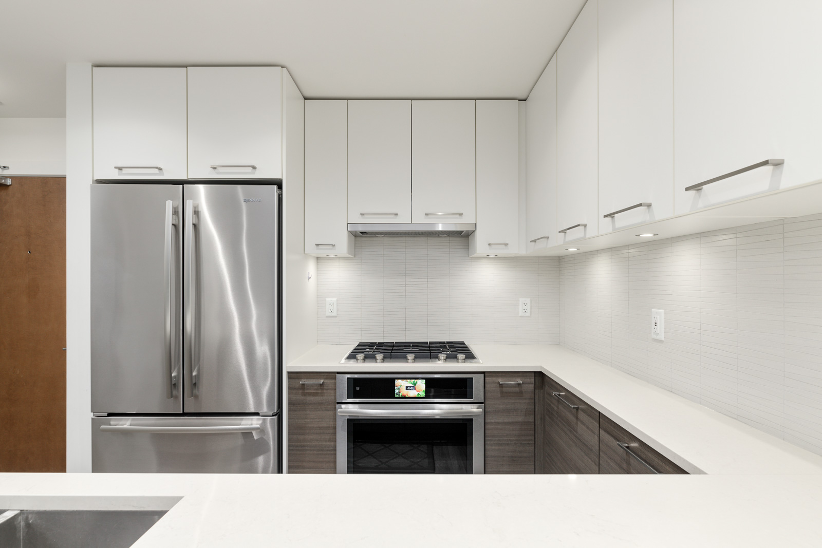 Modern kitchen with white cabinets, stainless steel refrigerator, stove, and oven. The countertops are white, and the backsplash is light grey tile.