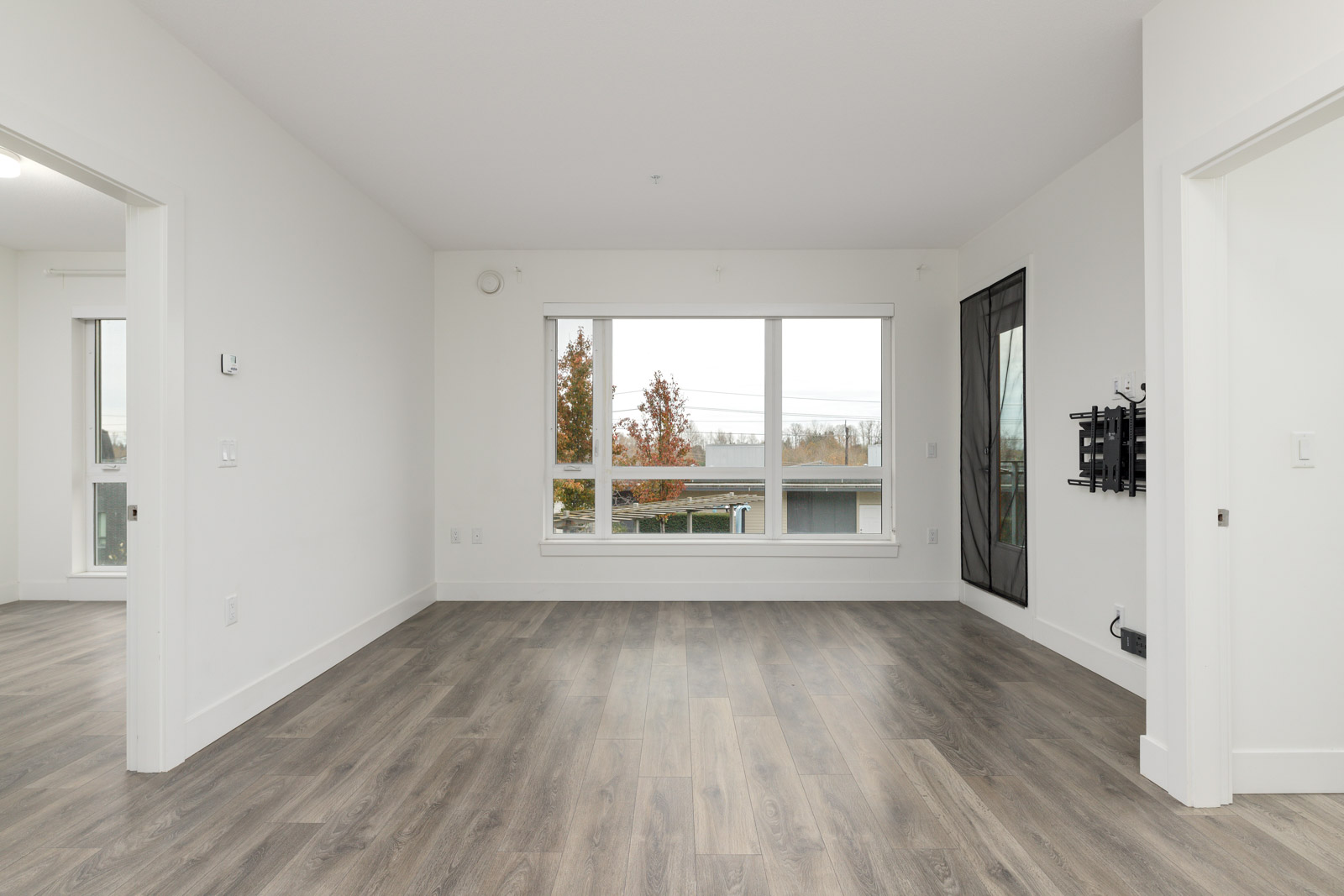 Empty room with large windows, white walls, and wood flooring. Wall-mounted TV bracket is visible. Two open doorways are on each side.