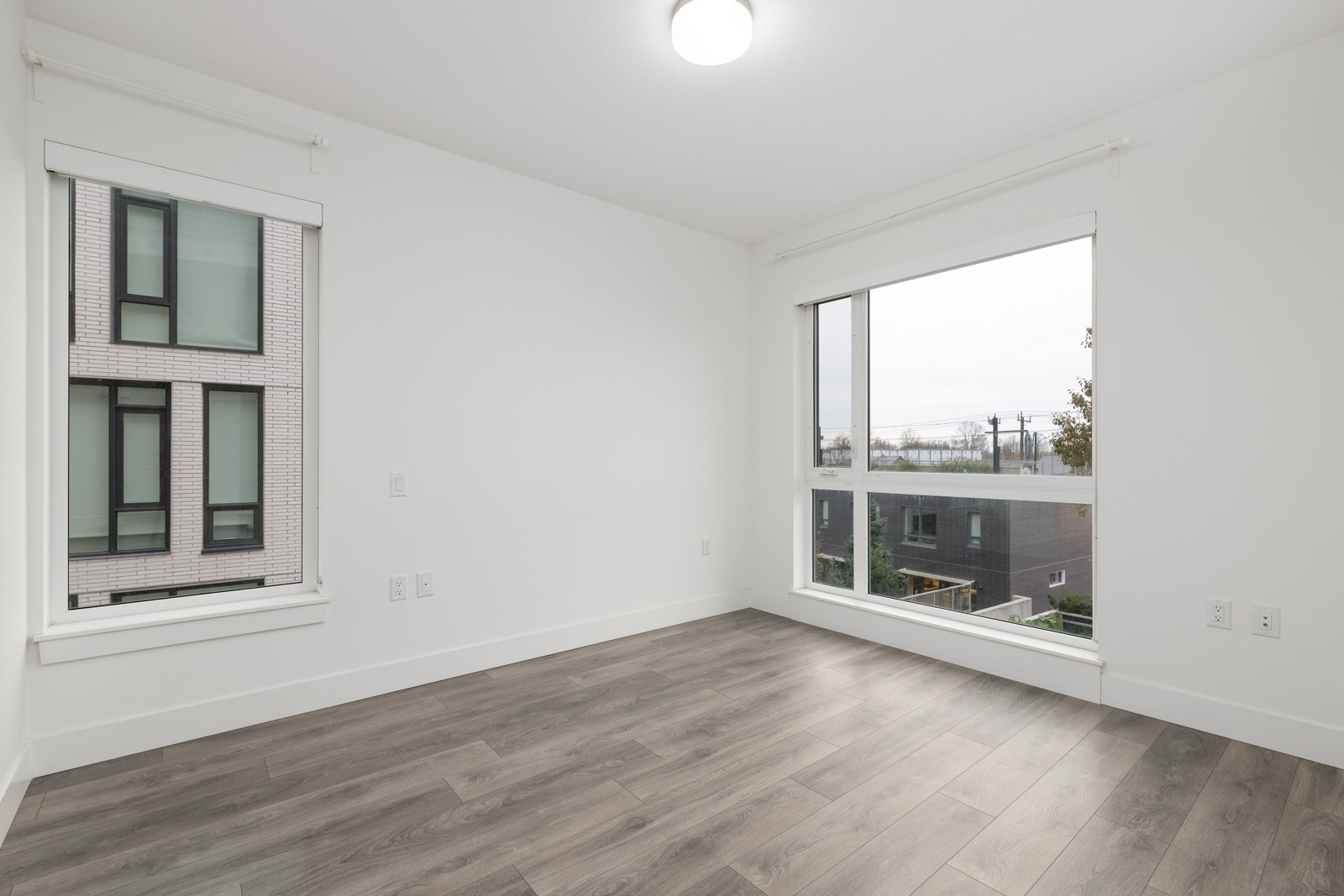 Empty room with light gray walls, wooden floor, and two large windows overlooking modern buildings and a partially visible landscape outside.
