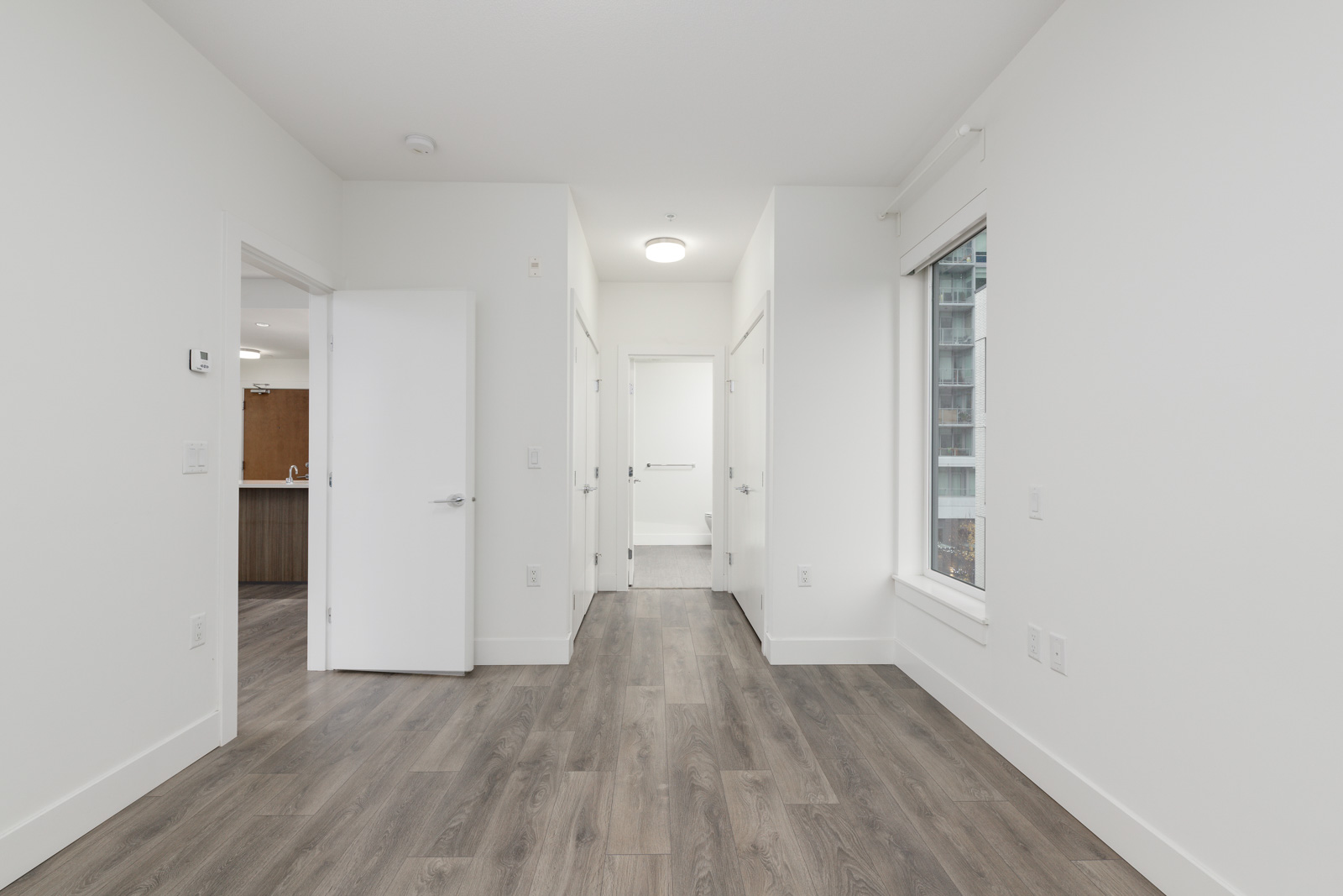 Empty room with white walls, wood flooring, an open door to a kitchen, and a large window.