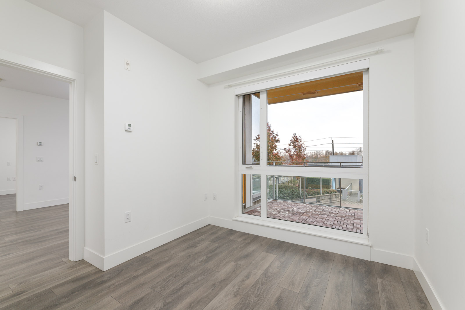 Empty room with a large window revealing a balcony and trees outside. The room has white walls and wood flooring.