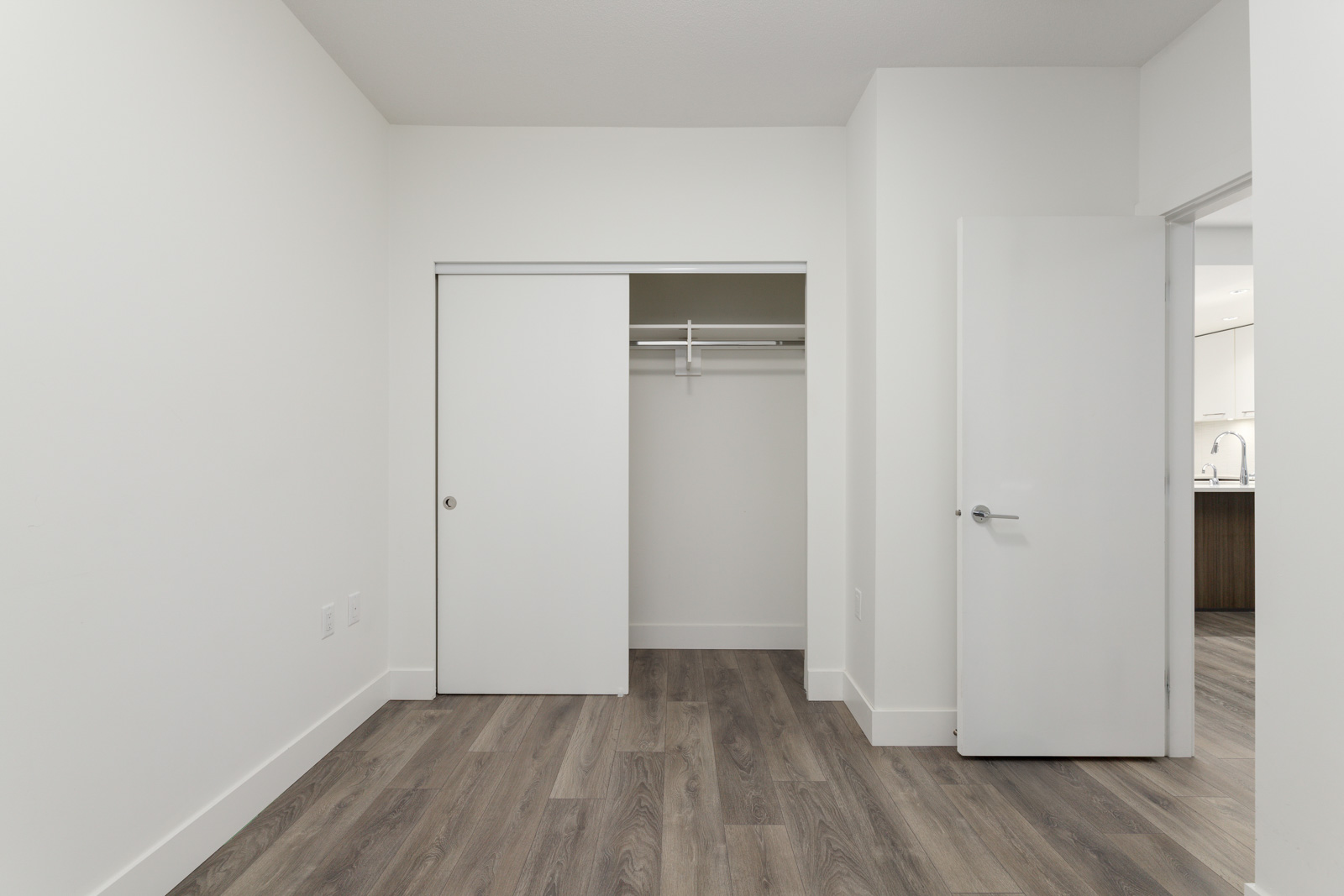 Empty room with light gray walls, wood flooring, an open closet with sliding doors, and a white door leading to another room.
