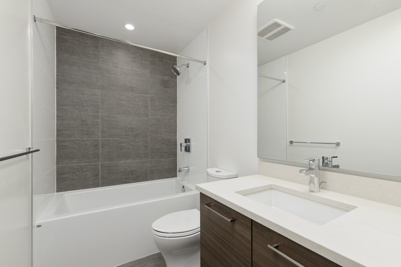 Modern bathroom with white fixtures, a bathtub with a shower, dark tiled wall, large mirror, and wooden vanity with a white countertop.