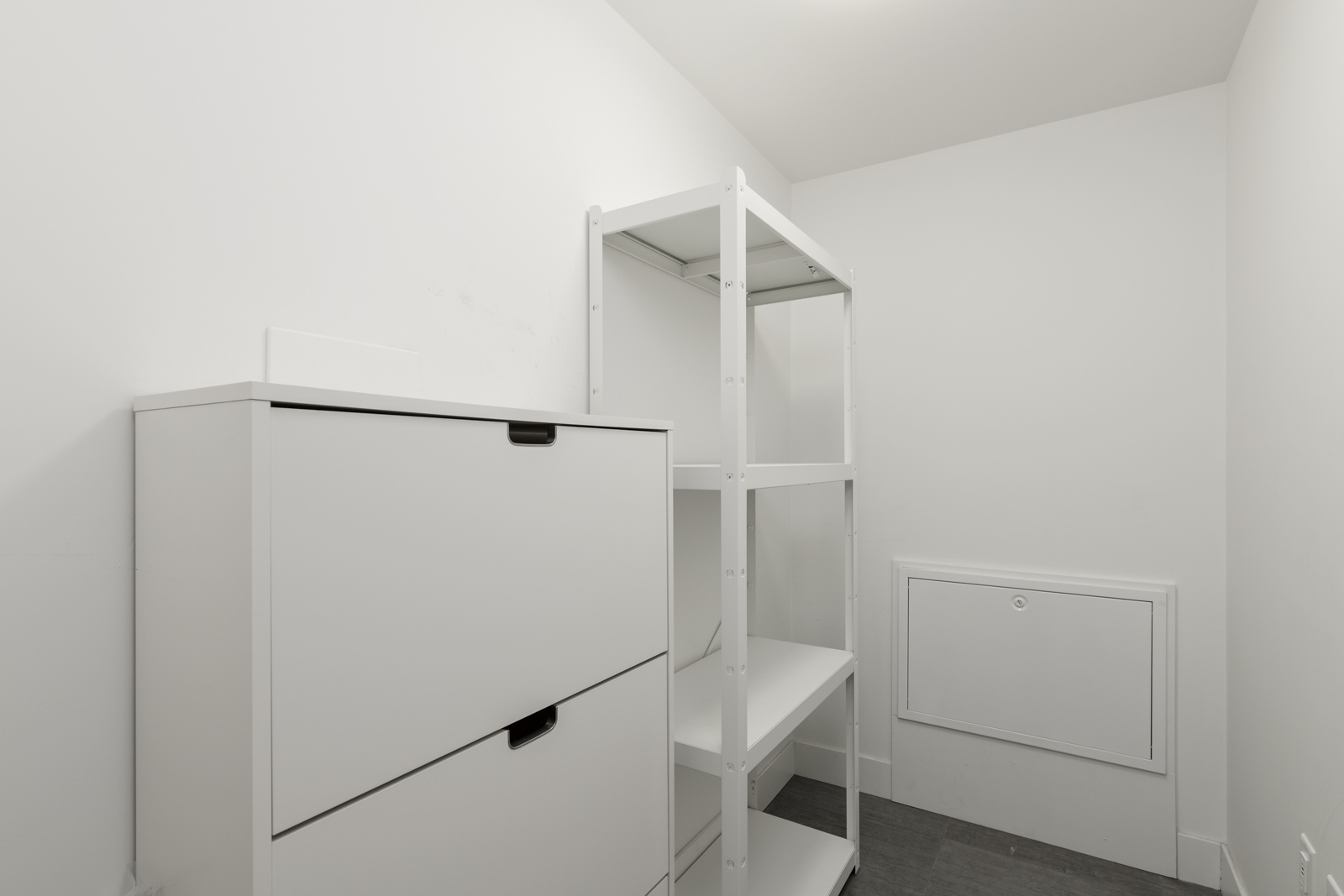 A small, white storage room with a shelving unit and a cabinet, featuring a utility hatch on the back wall.