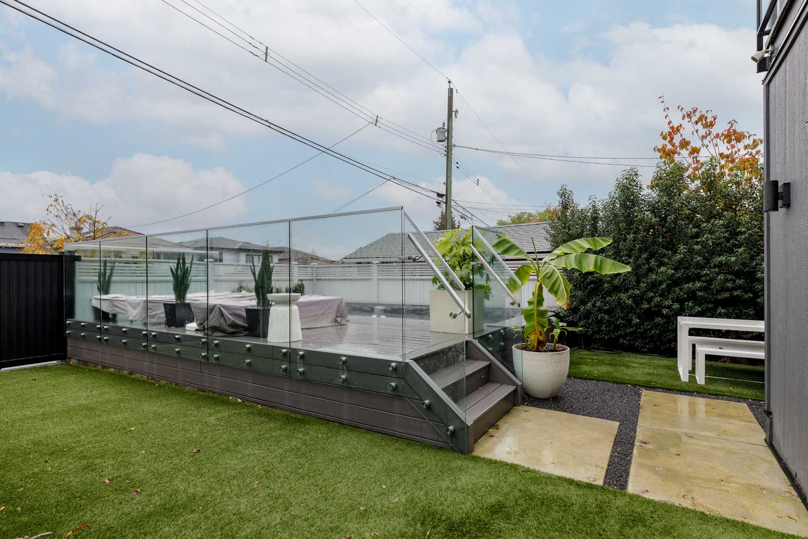 A modern backyard with a glass-enclosed outdoor seating area, potted plants, and a small lawn. A white bench is in the background, and the sky is cloudy.
