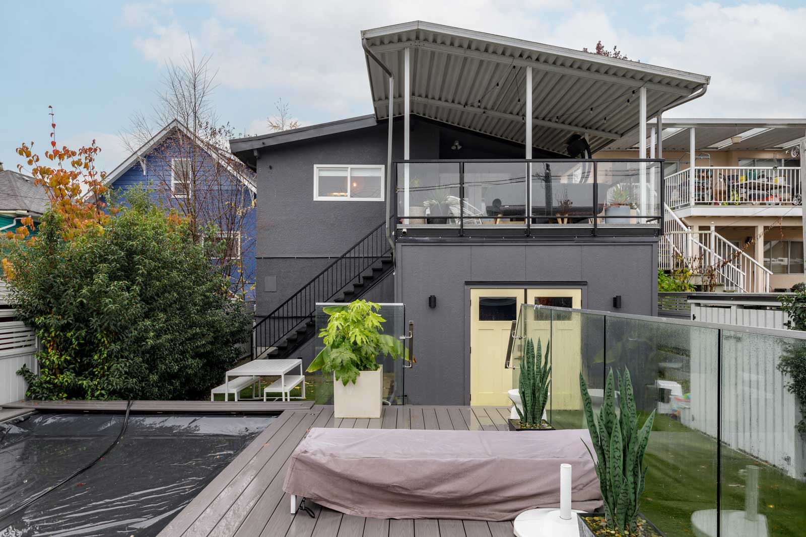 Modern two-story house with a covered balcony, outdoor seating, potted plants, and a rectangular pool on a wooden deck. Nearby, a blue house is partially visible. Overcast sky in the background.