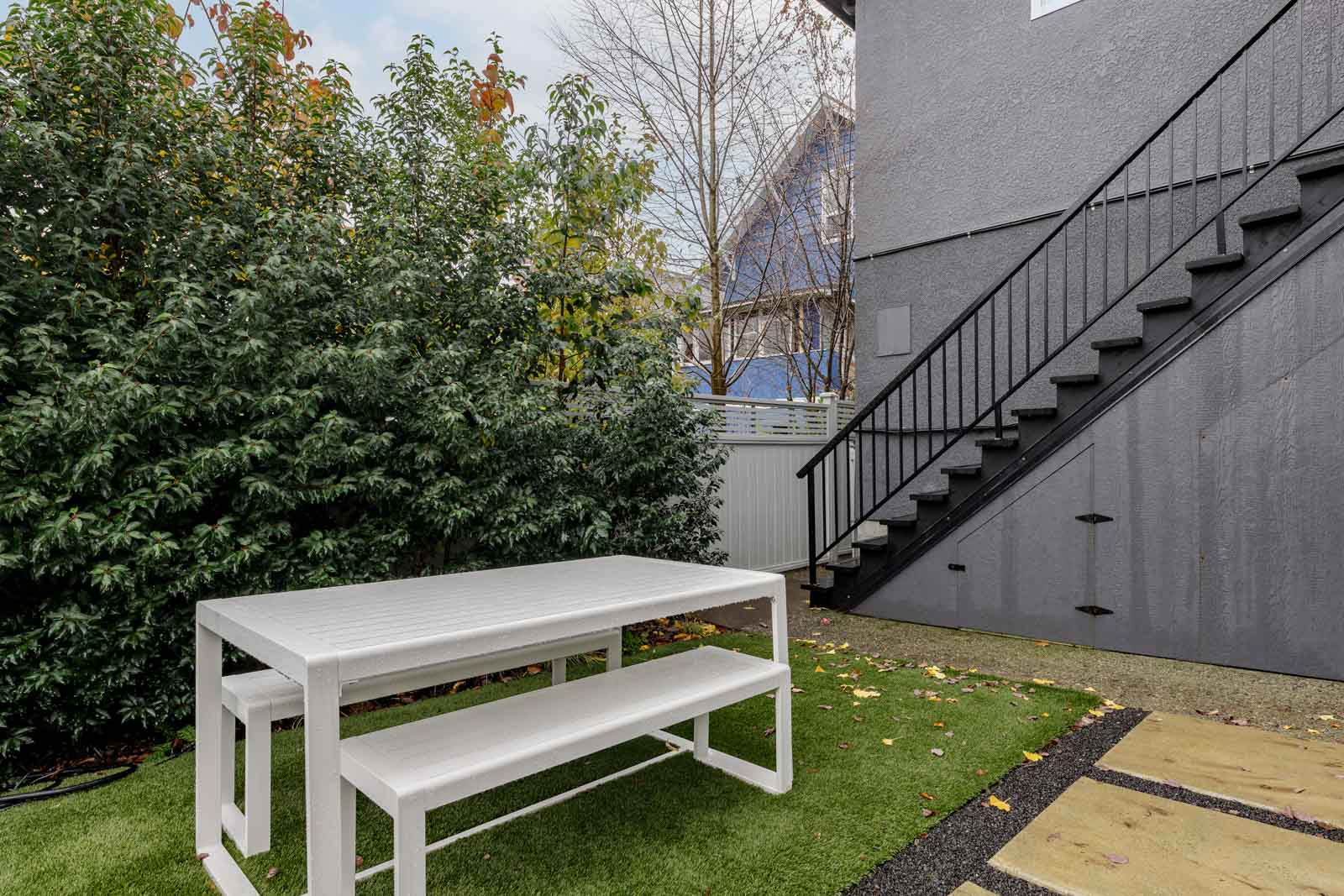 Outdoor area with a white picnic table and benches on a patch of grass, next to a gray building with a black metal staircase.