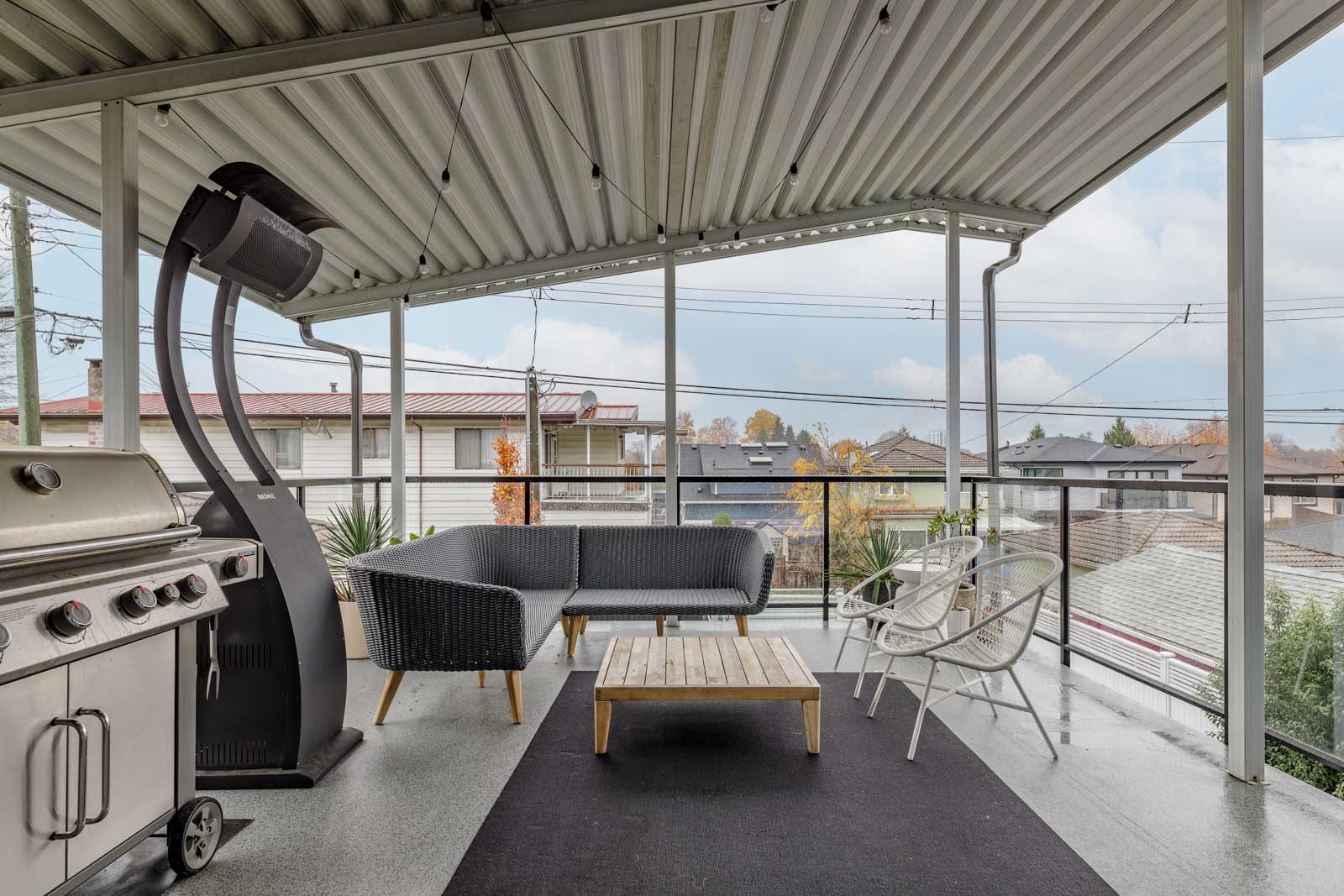 Covered patio with a barbecue grill, black sofa, wooden coffee table, two white chairs, and a space heater. Overlooks a residential neighborhood.