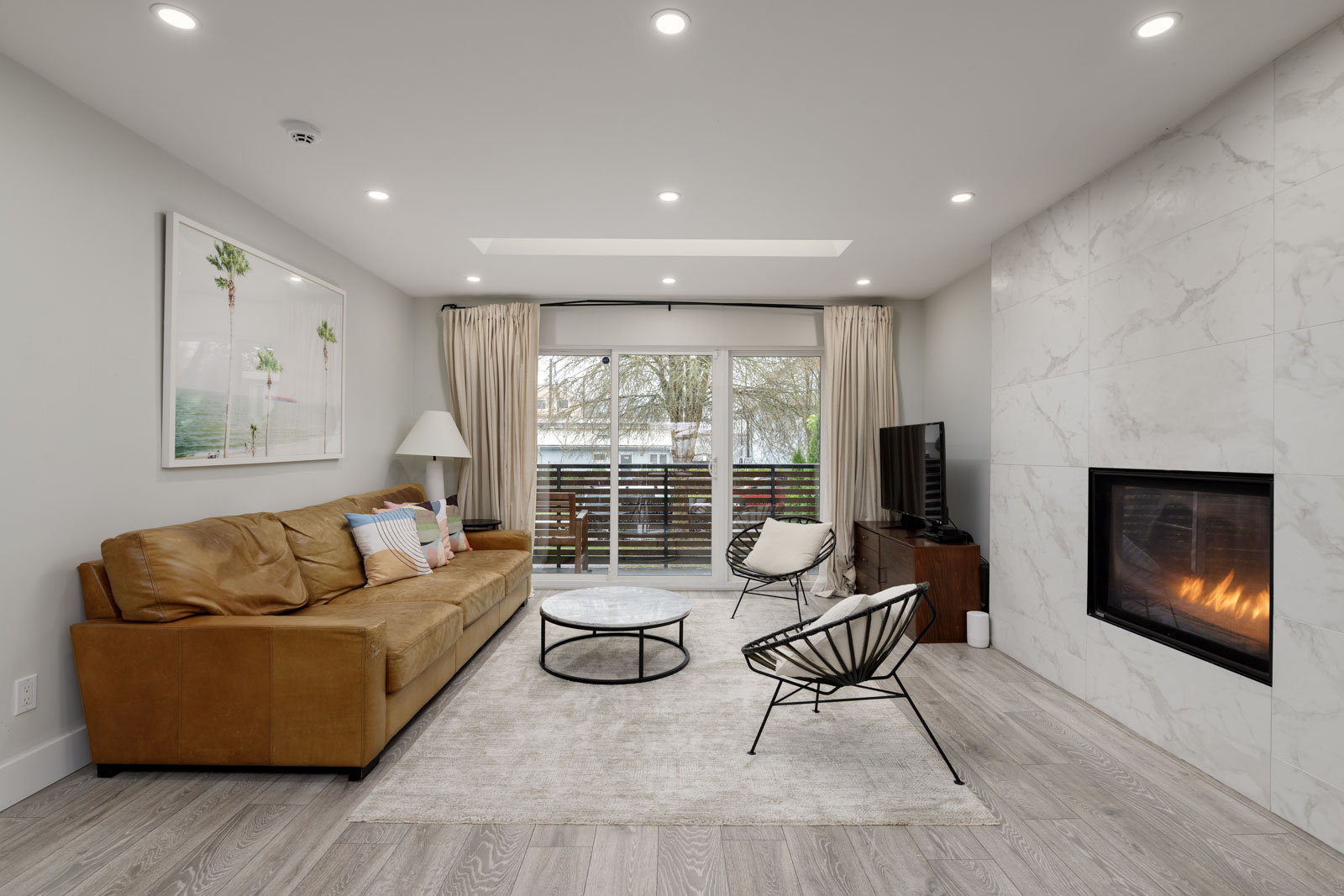 A modern living room with a tan sofa, armchair, marble fireplace, and wall-mounted TV. Large windows with curtains offer a view of trees outside. A round coffee table sits on a neutral rug.