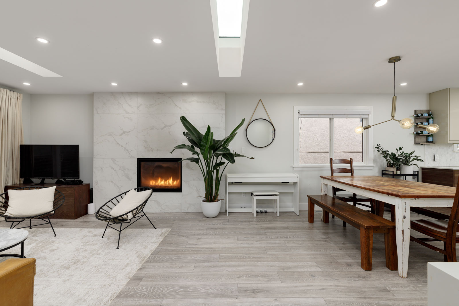 Modern living and dining area with wood furniture, a large plant, a wall-mounted circular mirror, and a fireplace. A window brings in natural light, complementing the light-colored flooring and decor.