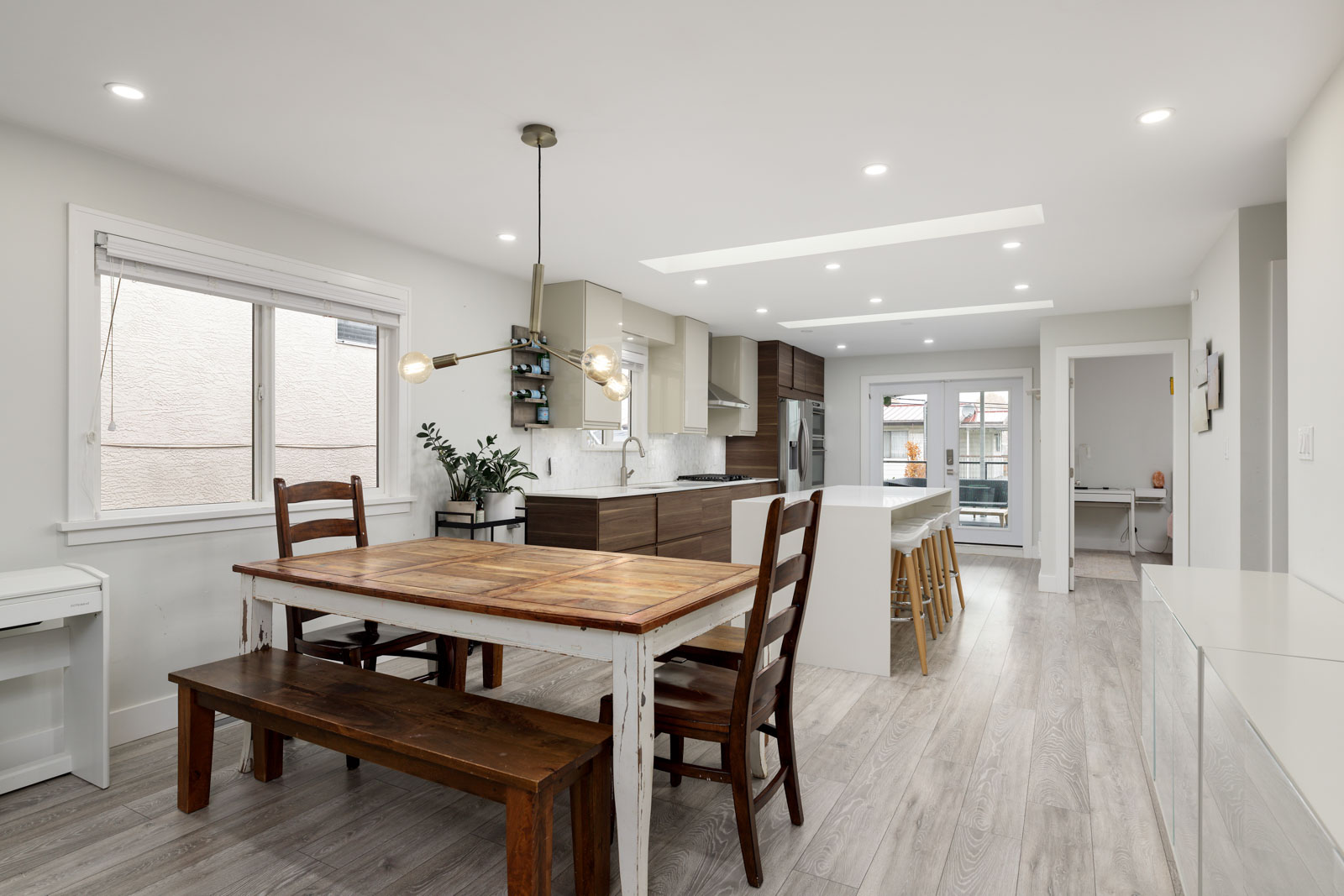 Modern kitchen and dining area with wooden table, chairs, and bench. Features an island with bar stools, hanging light fixture, and contemporary decor. Natural light from windows.