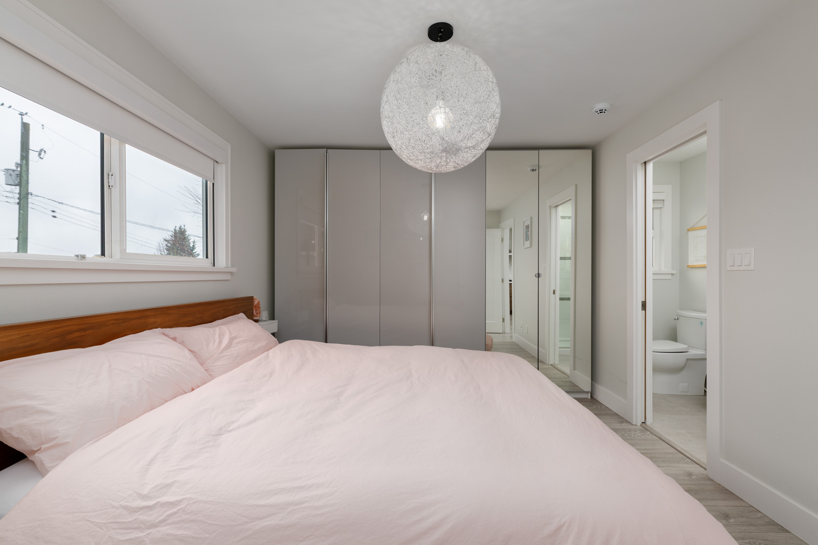 A bedroom with a large bed, pale pink bedding, a spherical ceiling light, a wardrobe with mirrored doors, and an attached bathroom.