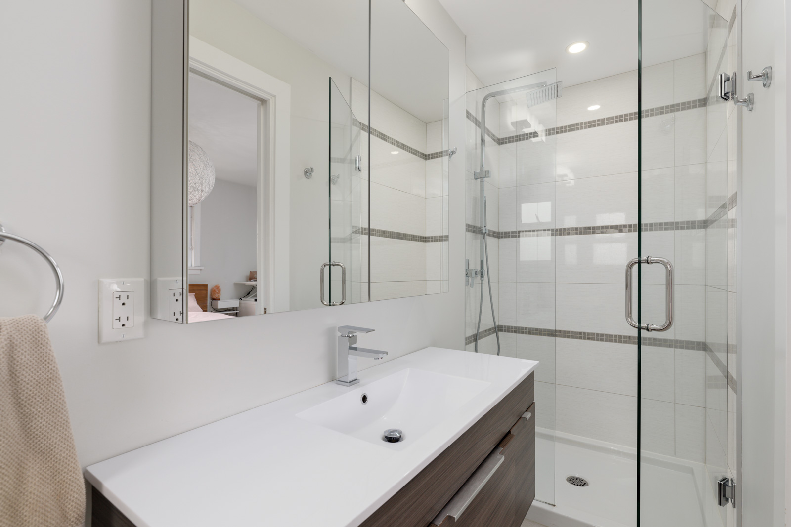Modern bathroom with glass shower, white sink, and wooden vanity. Neutral walls, towel on rack, and a glimpse of an adjacent room through a small window.