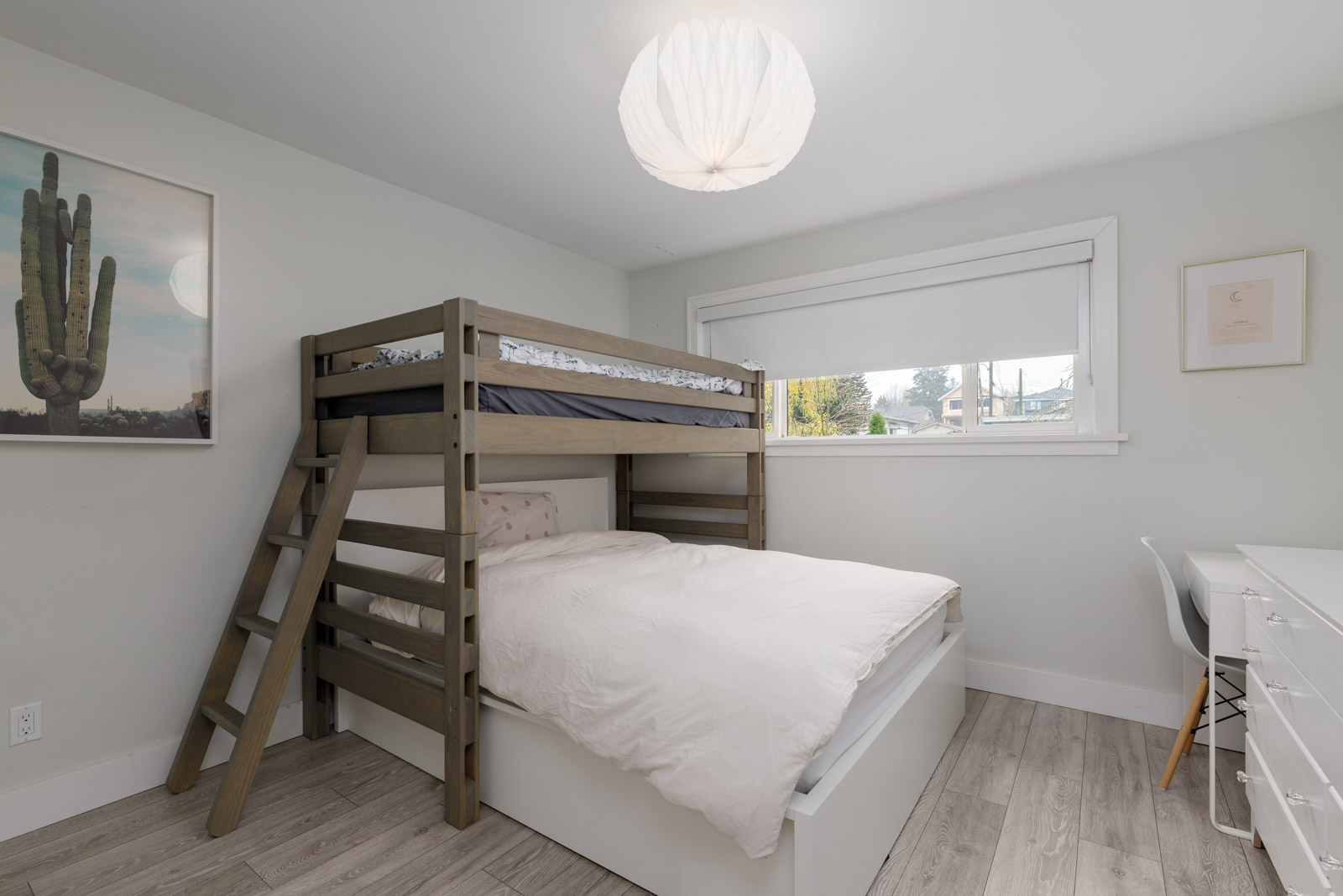 Room with a bunk bed, light walls, wood floors, cactus art, a large window, and a desk with a chair.