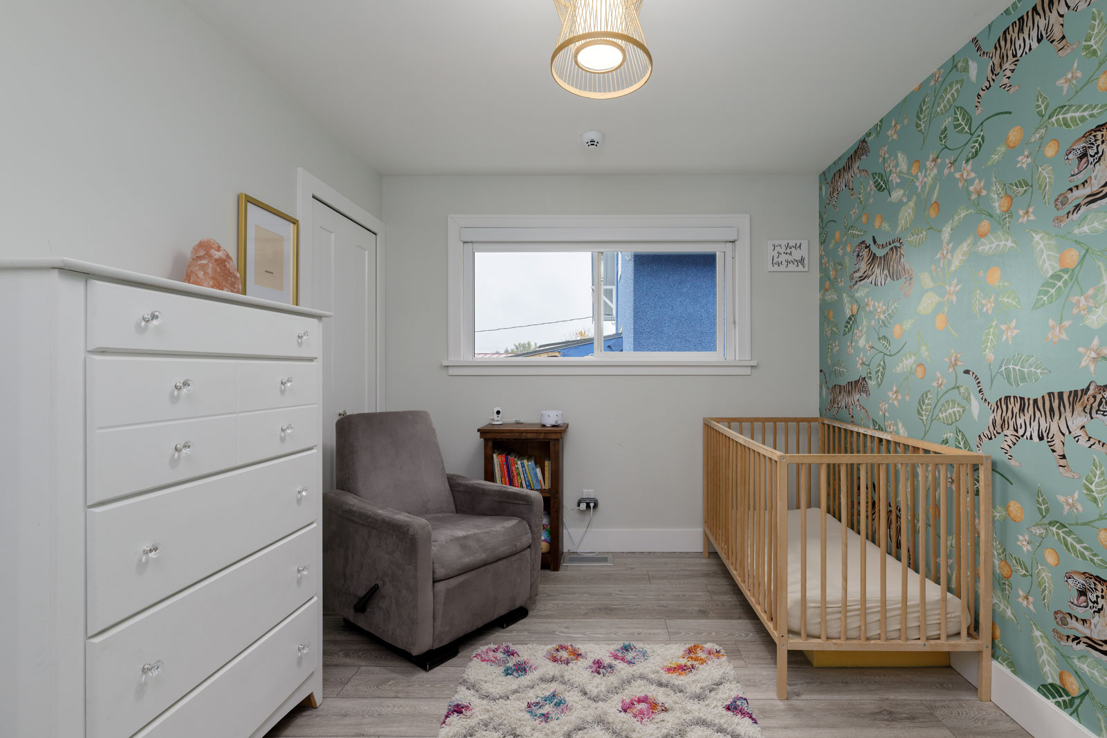 A nursery with a white dresser, gray armchair, wooden crib, and colorful wall with jungle-themed wallpaper. A window and a small bookshelf are also visible.