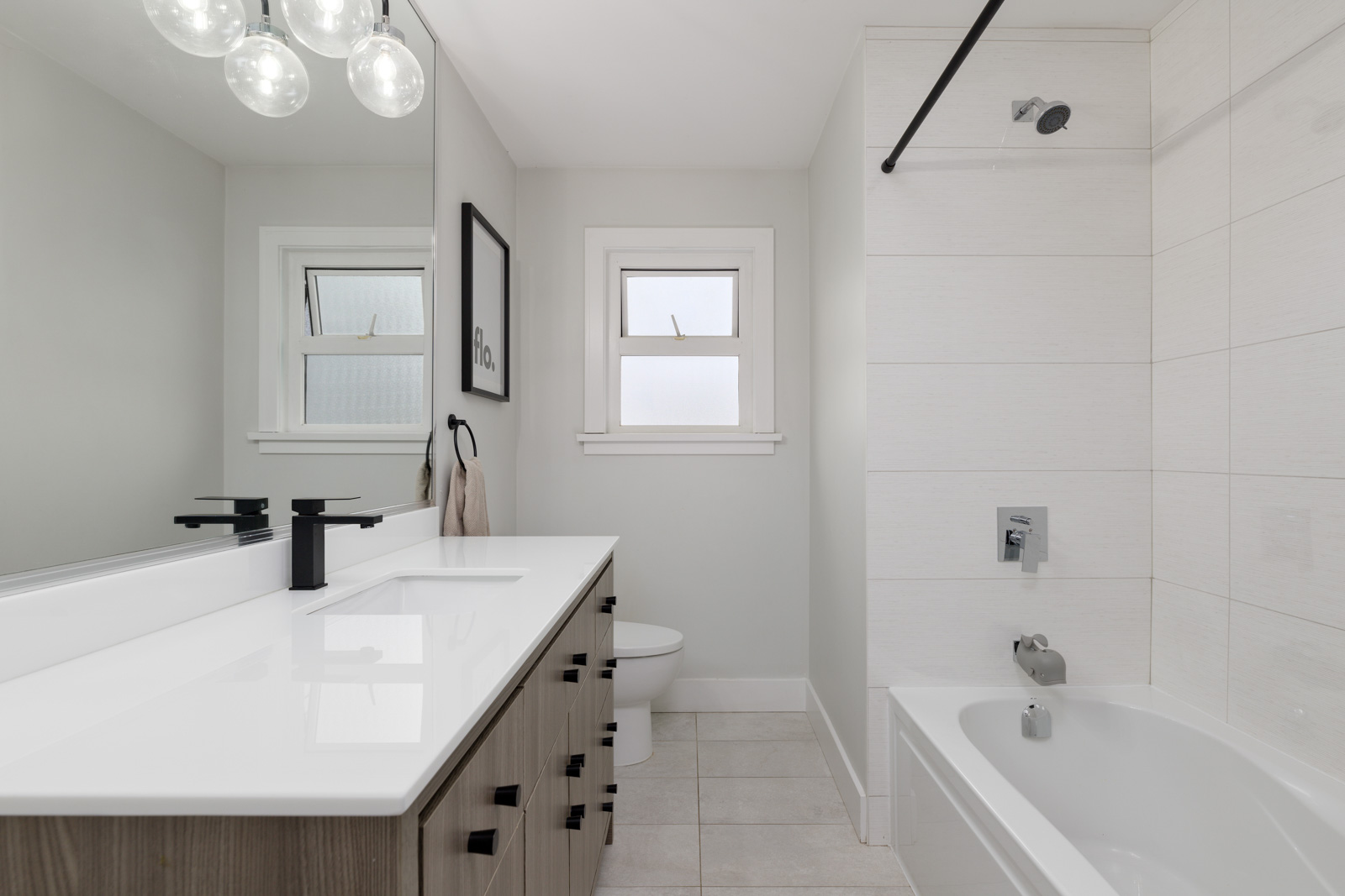 Modern bathroom with a double sink vanity, black faucets, bathtub with shower, large mirror, wall art, and two frosted windows.