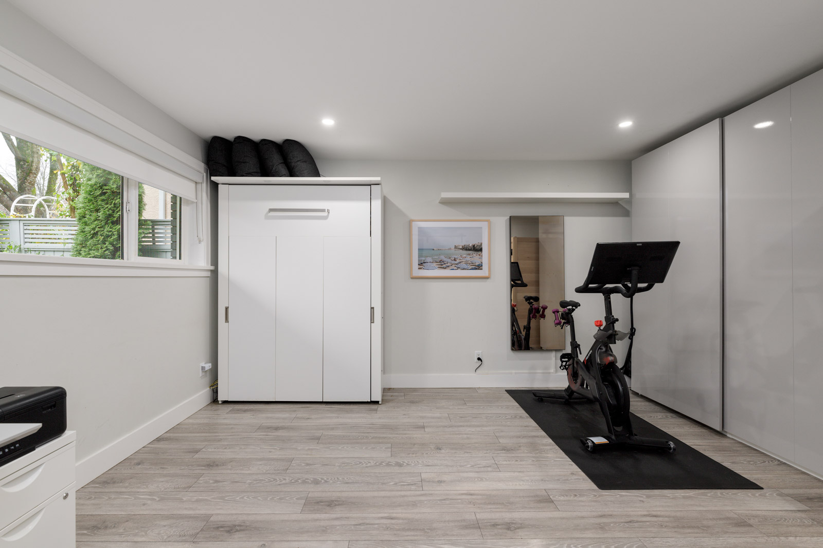 Home gym with stationary bike on a mat, large mirrors, and built-in storage cabinets. Bright space with wooden flooring and natural light from a window.