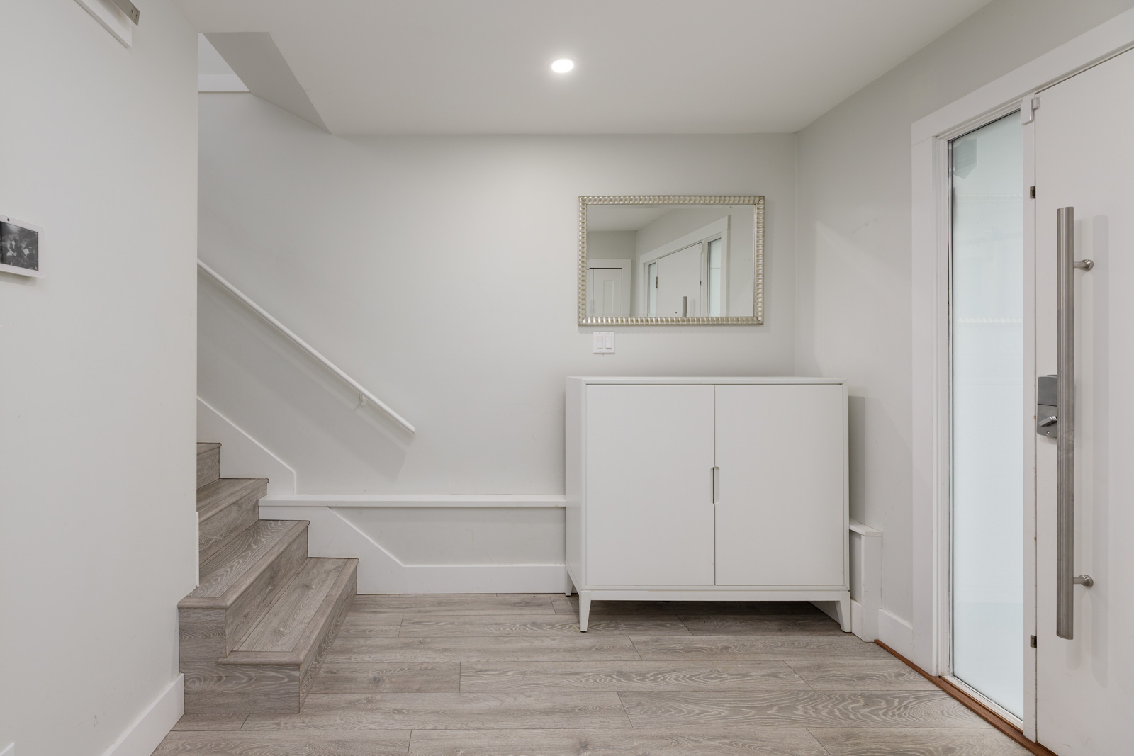 Minimalist hallway with a white cabinet, mirror above, wooden stairs on the left, and a glass door on the right. Neutral tones and recessed lighting complete the space.