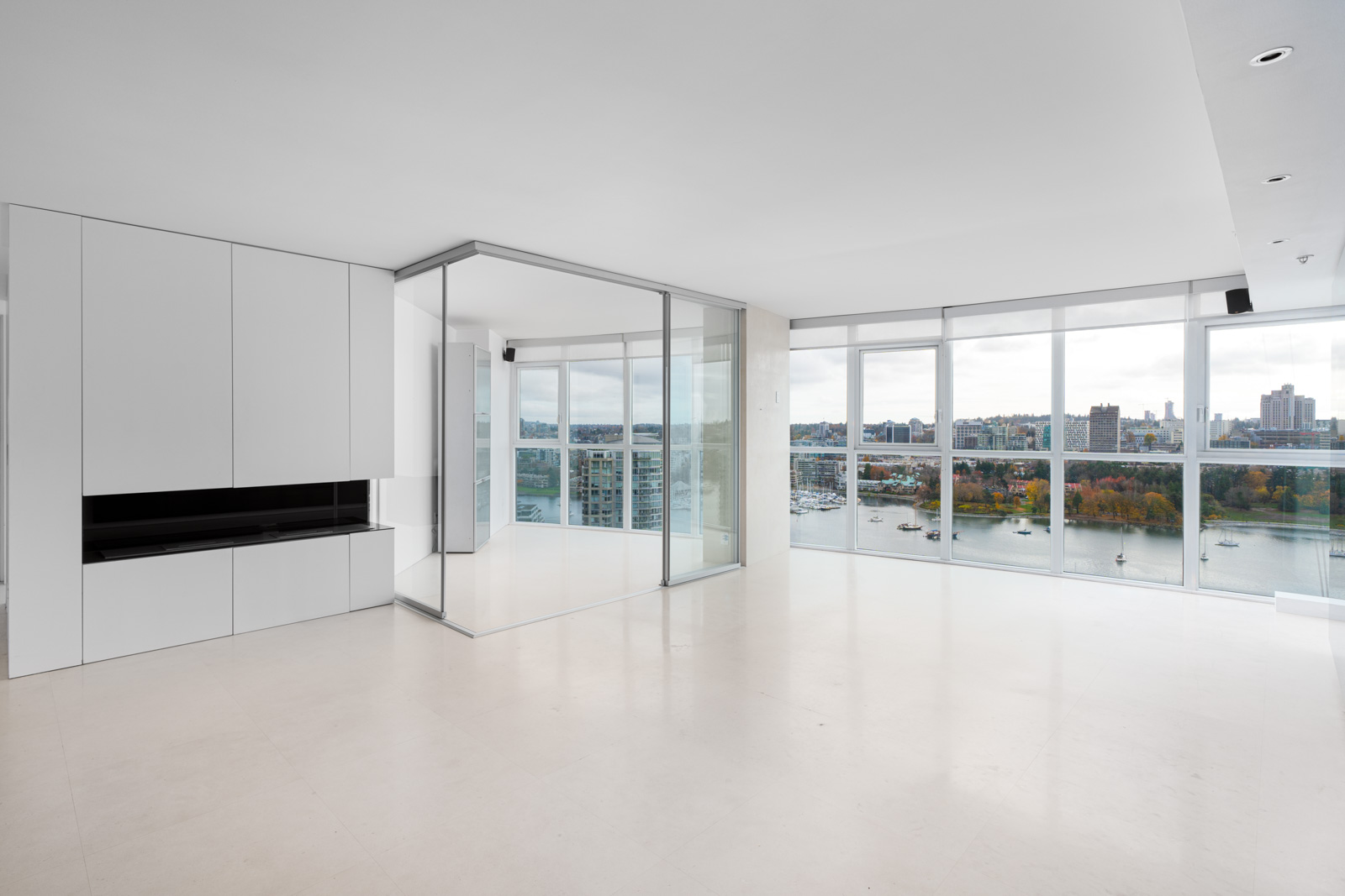 Modern, empty apartment with large windows, white walls, minimalist cabinetry, and a view of a river and cityscape.