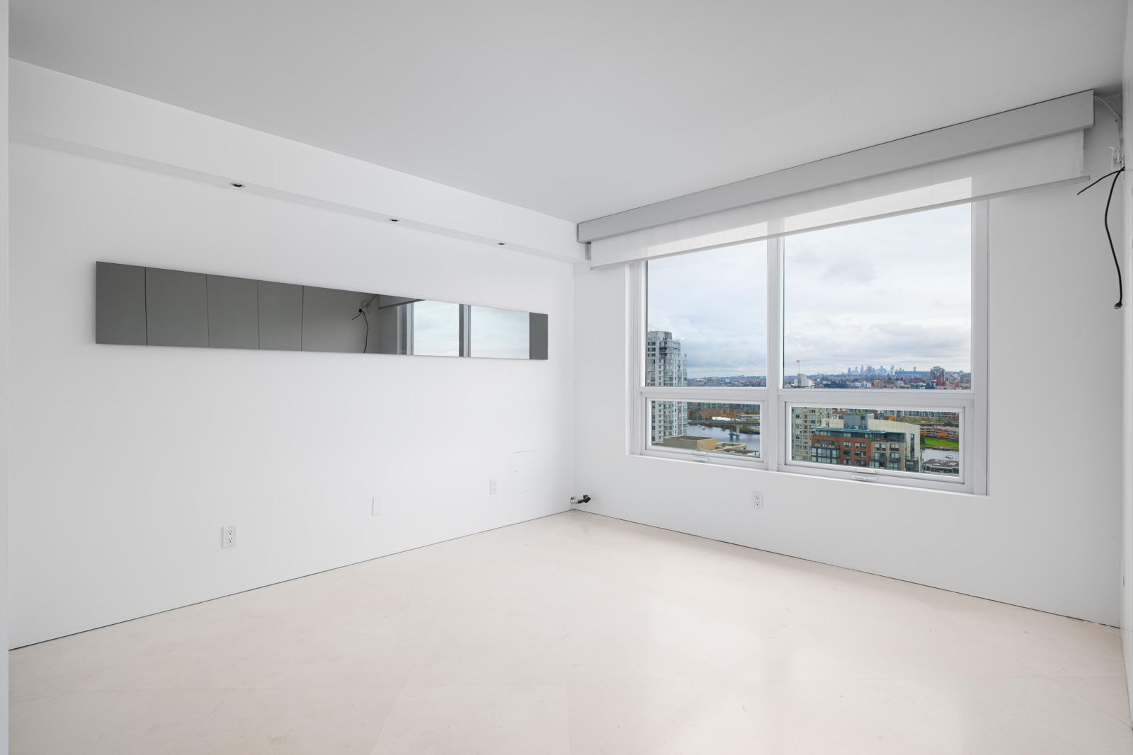Empty white room with large window featuring city view. Wall mirror on the left side.