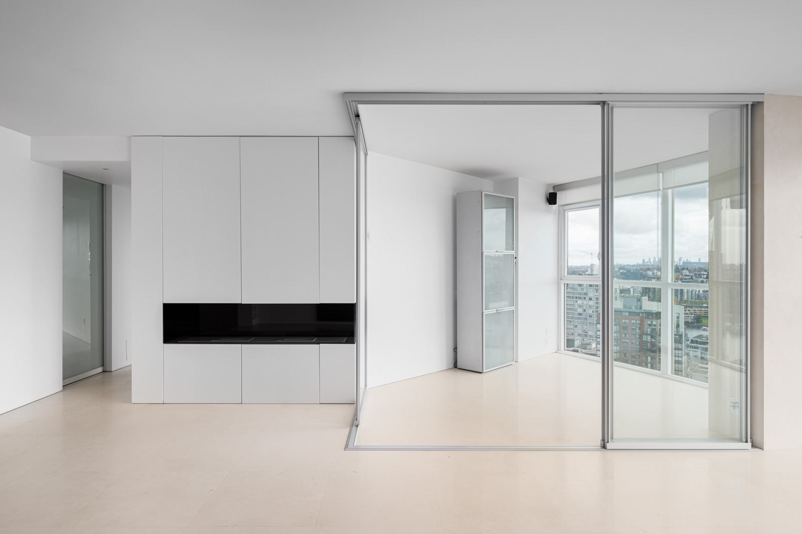 Modern, minimalistic interior with white walls, sliding glass doors, and built-in storage. Floor-to-ceiling windows offer a cityscape view.