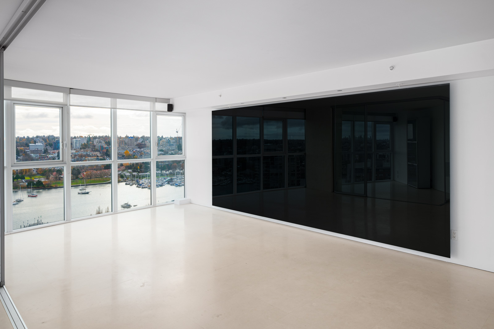 Spacious empty room with large windows overlooking a cityscape and waterfront, featuring white walls and a light-colored floor.