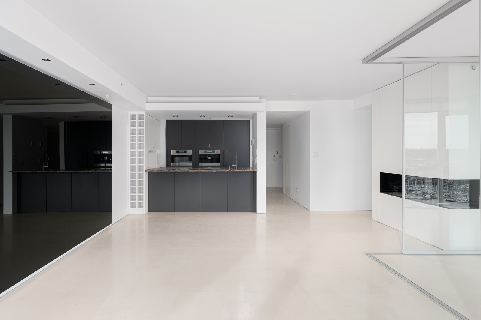 A modern, minimalist kitchen with dark cabinetry, built-in ovens, and a light floor. The room has a glass partition and reflects a spacious, open design.