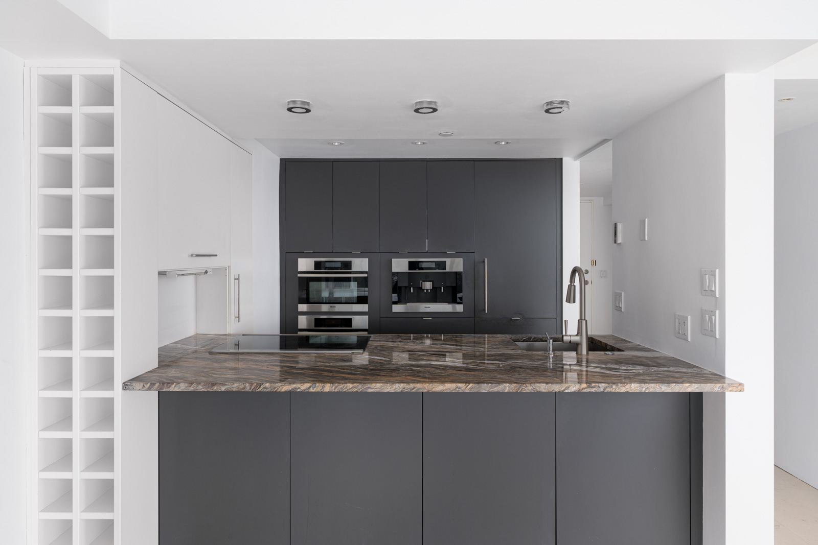 Modern kitchen with dark cabinets, marbled countertop, built-in ovens, and a wine rack. White walls and ceiling lights complete the sleek design.