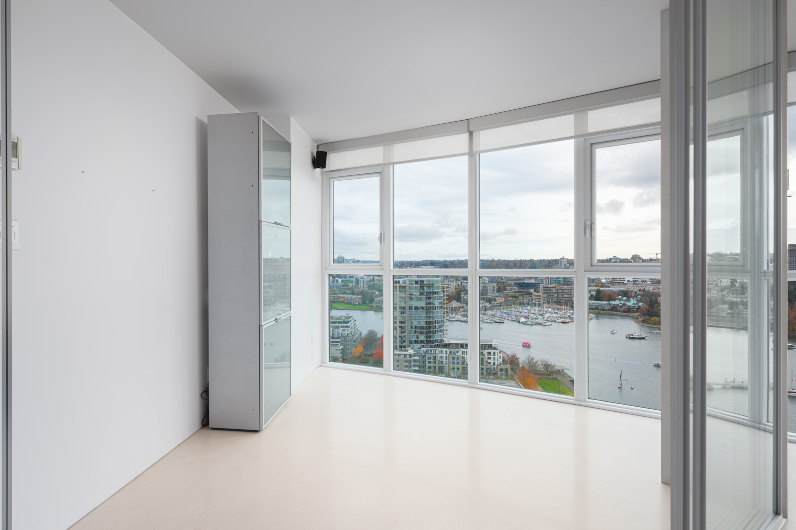 Empty room with white walls and a glass cabinet, featuring large windows offering a view of a cityscape and waterfront with boats.