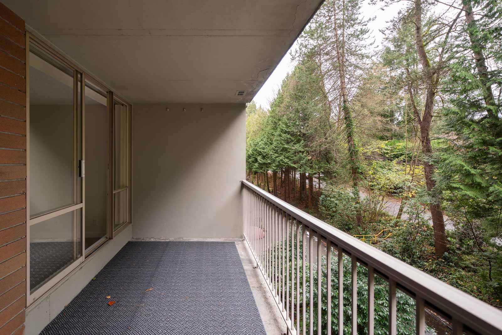 A balcony with a metal railing overlooks a wooded area. There is a glass sliding door leading inside, and the floor has a textured mat.