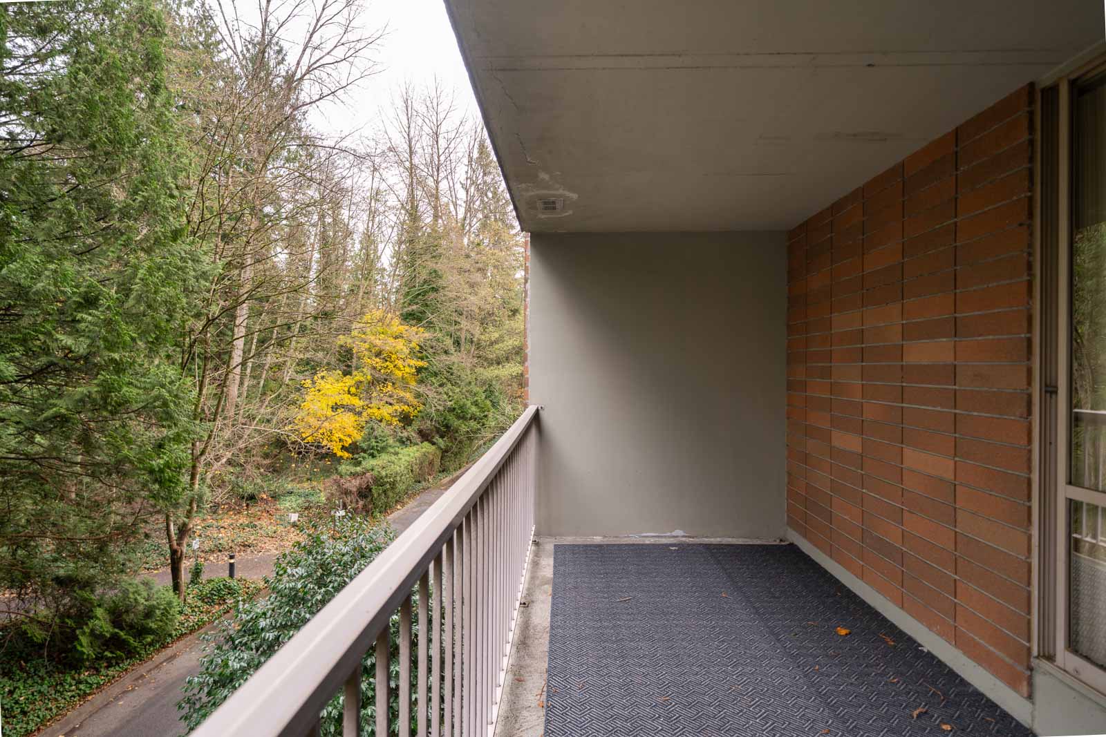 Balcony view with brick wall, railing, and a rubber mat. Overlooks a wooded area with trees, some with autumn leaves.