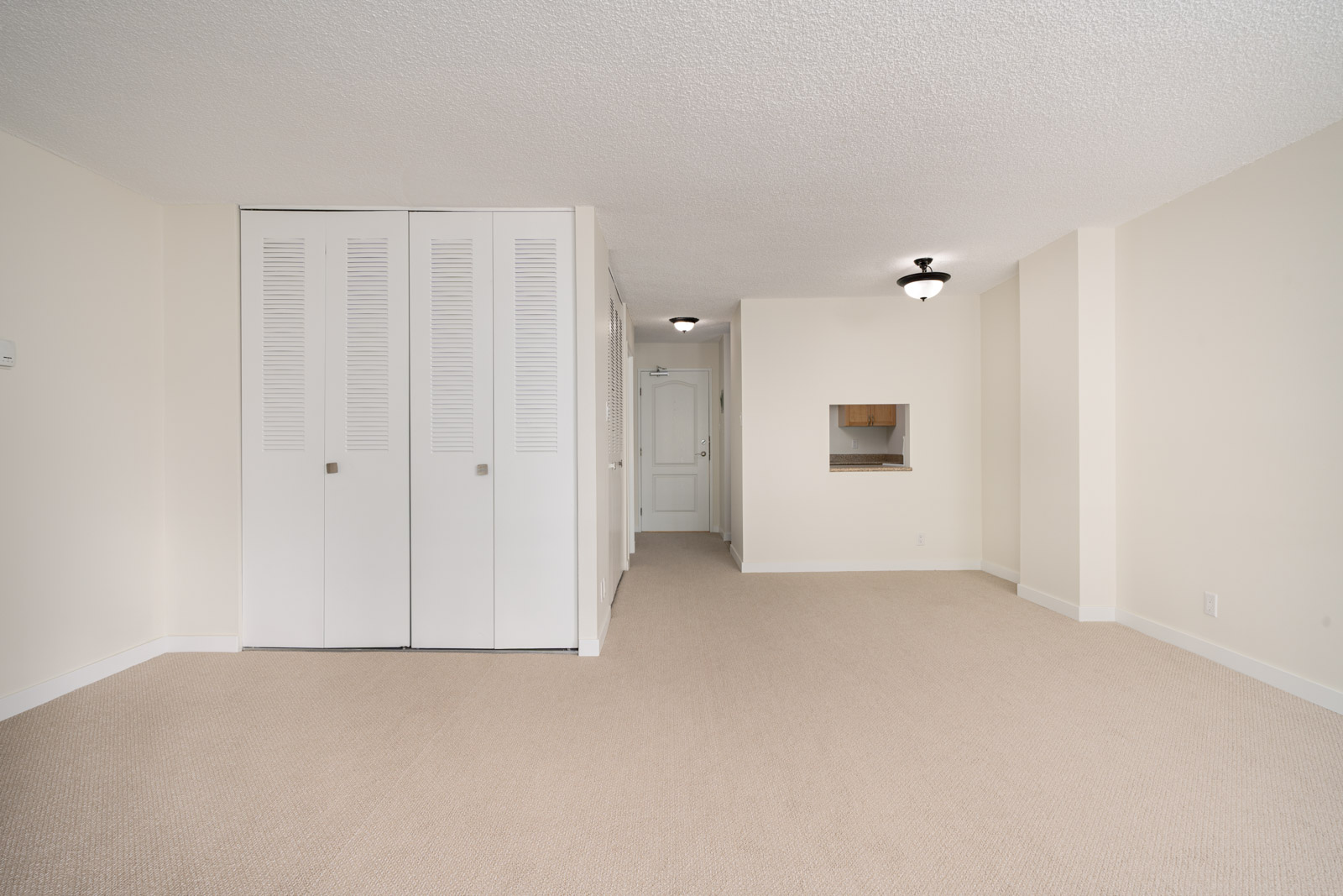 Empty room with beige carpet, white walls, and closet with louvered doors. Features a recessed light fixture and a small pass-through window to another area.