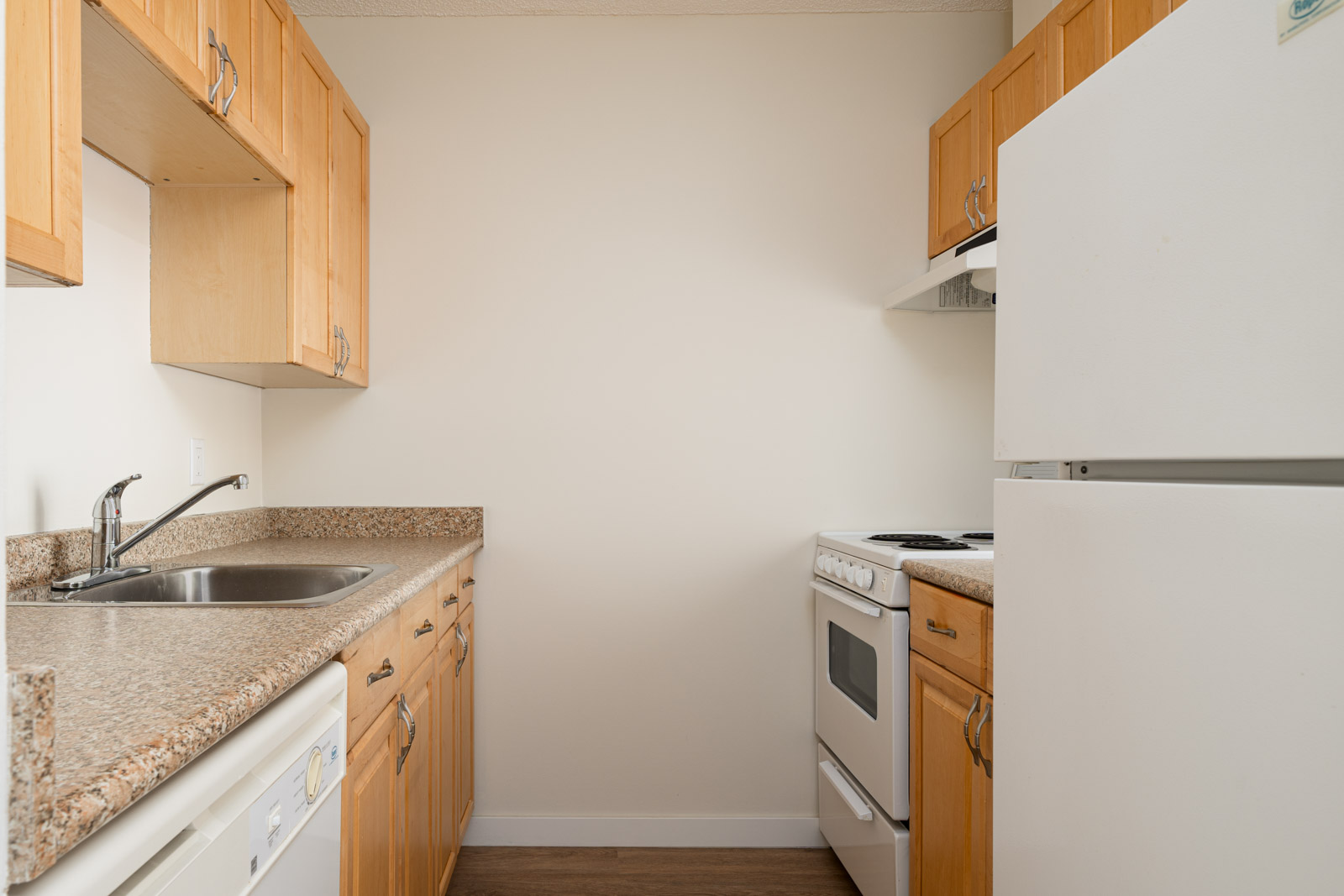 A narrow kitchen with light wood cabinets, granite countertops, a white stove, dishwasher, and refrigerator against white walls with a wooden floor.