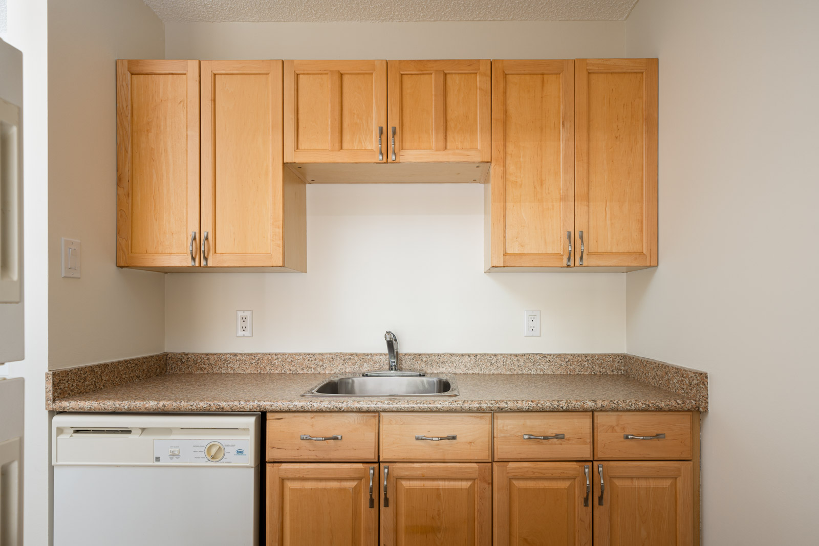 A small kitchen with wooden cabinets, granite countertops, a stainless steel sink, and a built-in dishwasher.