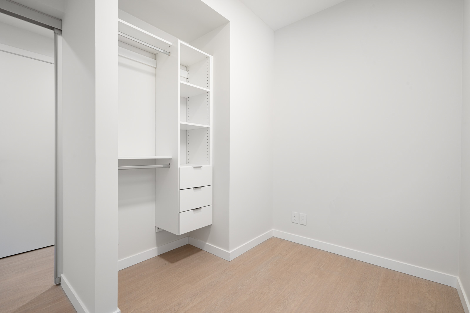 Empty white closet with built-in shelves and hanging rod, featuring light wood flooring.
