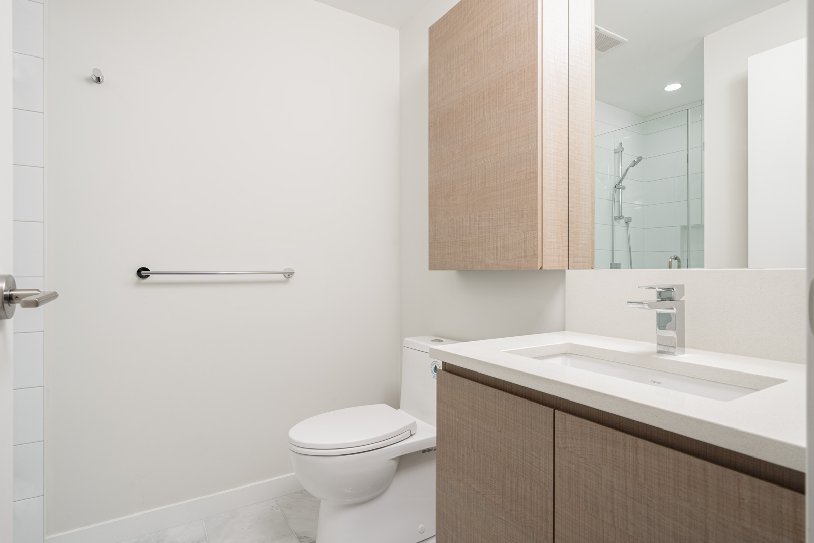 Modern bathroom with a white toilet, wooden cabinets, a countertop sink, and a wall-mounted towel bar. A glass shower enclosure is visible in the background.