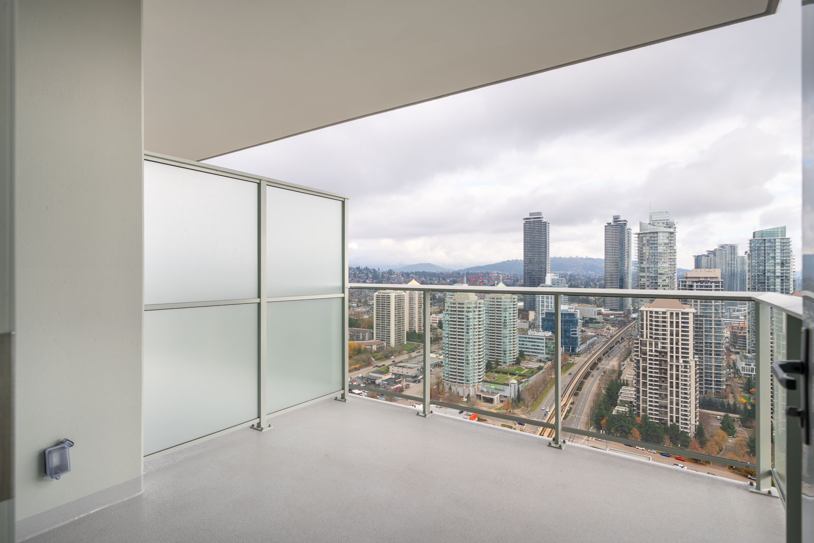 High-rise balcony overlooking a cityscape with tall buildings, roads, and cloudy skies. The balcony features glass railings and frosted privacy panels on one side.