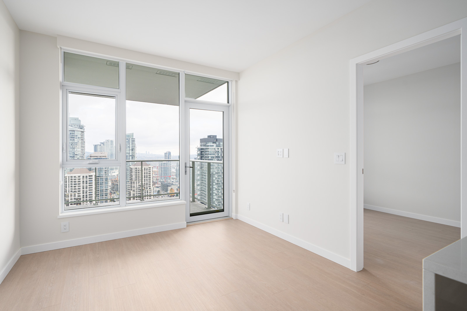 Bright, empty room with light wood flooring, large windows, and a sliding glass door leading to a balcony overlooking a cityscape.
