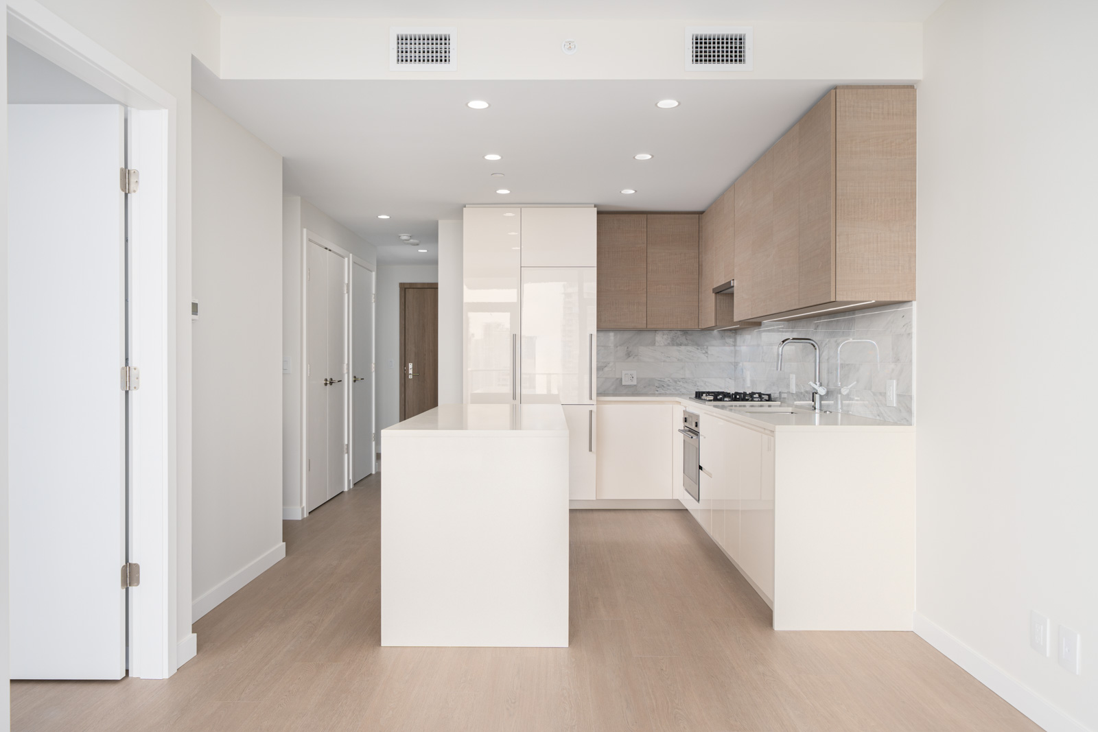 Modern kitchen with white island, wood cabinets, marble backsplash, and recessed lighting. Light wood flooring and a door to the left.