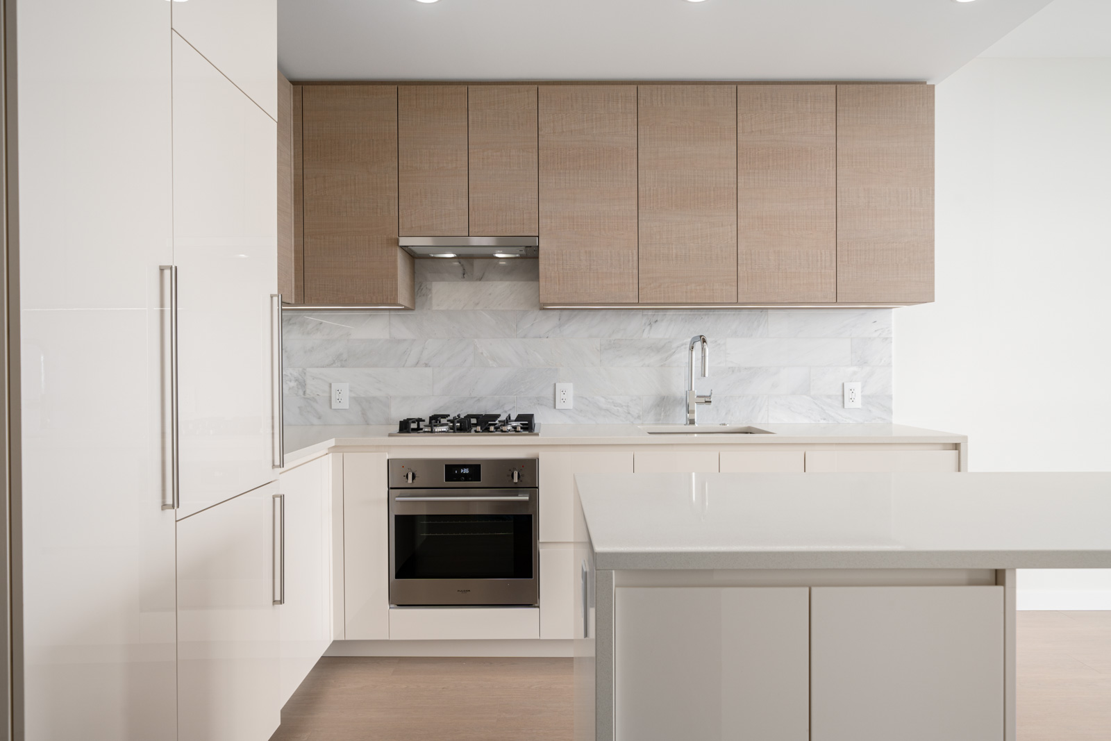 Modern kitchen with light wood cabinets, a marble backsplash, an oven with a cooktop, and an island with a glossy surface.