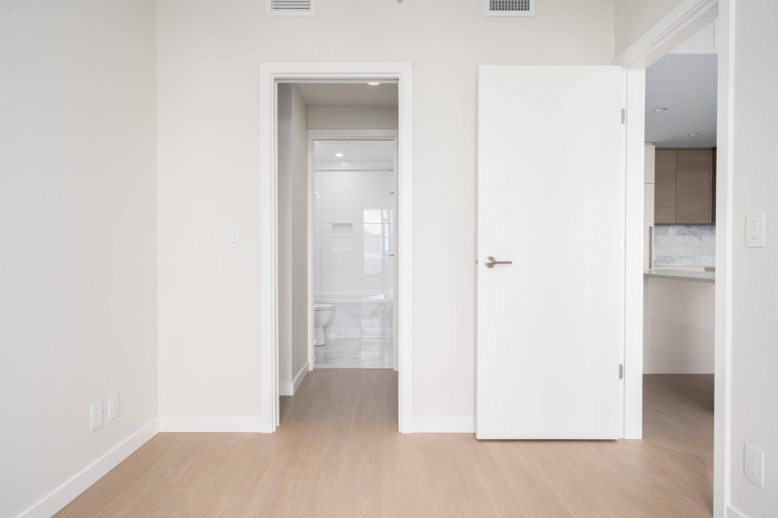 Minimalist room with light wood flooring, open door to a bathroom, and a hallway leading to a kitchen area. Walls and doors are white.