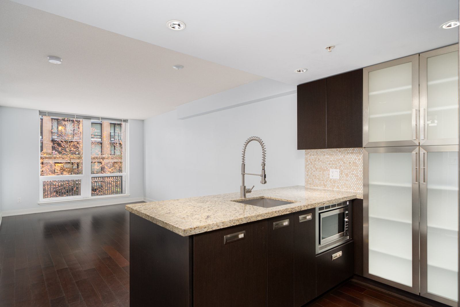 Modern kitchen with dark wood cabinets, granite countertop, and stainless steel appliances in an open living space with large window.