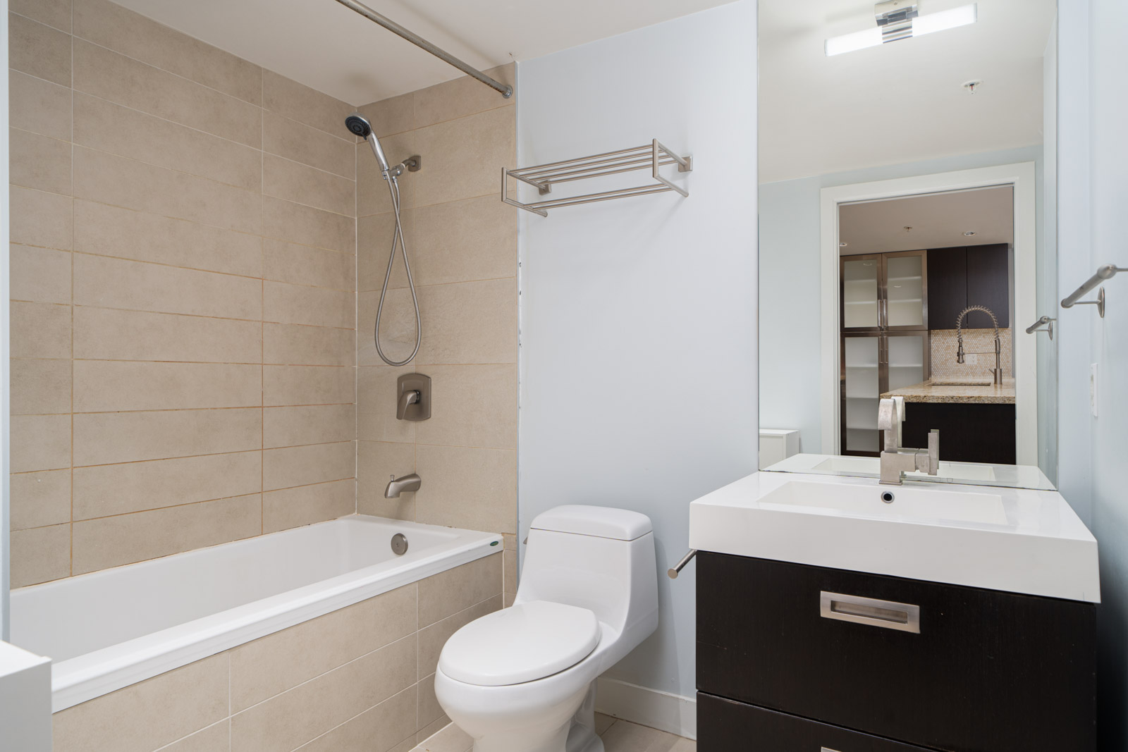 Modern bathroom with a bathtub, overhead shower, toilet, and rectangular sink with a mirror above. Light walls contrast with dark cabinetry.
